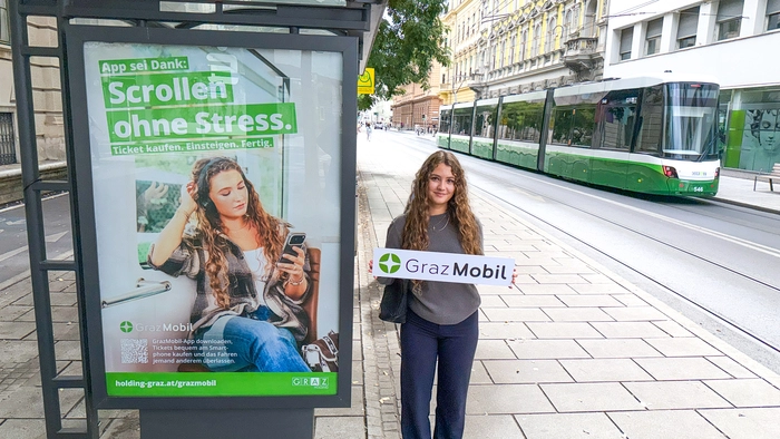 Eine junge Frau steht neben einer Haltestelle, auf der ein Sujet der Kampagne plakatiert ist. Sie hält ein Schild mit dem Logo der Graz Mobil-App.
