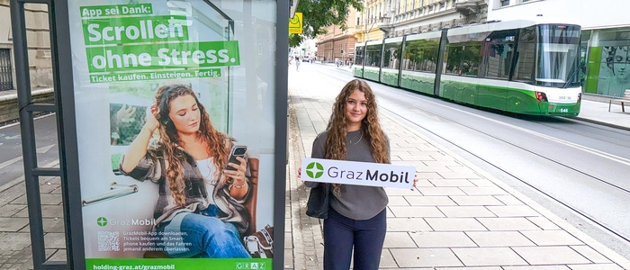 Eine junge Frau steht neben einer Haltestelle, auf der ein Sujet der Kampagne plakatiert ist. Sie hält ein Schild mit dem Logo der Graz Mobil-App.