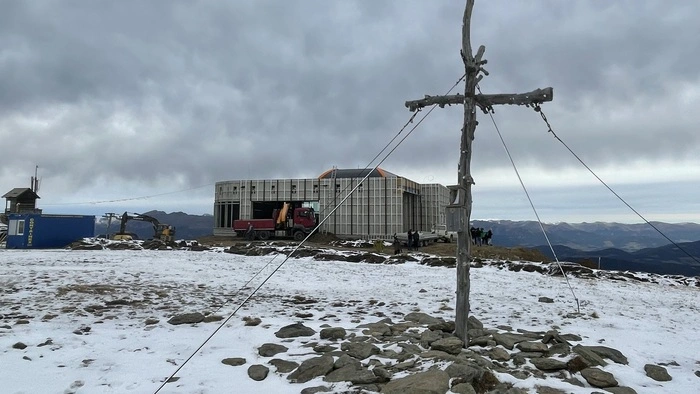 Ein Gipfelkreuz auf einem Berg, auf dem ein wenig Schnee liegt. Dahinter steht ein Gebäude, das noch nicht fertig errichtet ist. Um das Gebäude stehen Baumaschinen