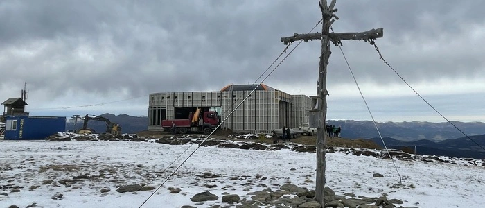 Ein Gipfelkreuz auf einem Berg, auf dem ein wenig Schnee liegt. Dahinter steht ein Gebäude, das noch nicht fertig errichtet ist. Um das Gebäude stehen Baumaschinen