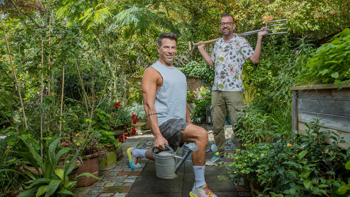 Das Bild zeigt Biogärtner Karl Ploberger und Fitnesstrainer Toni Klein in einem üppig grünen Garten mit vielen Pflanzen, Blumentöpfen und einem gepflasterten Weg. Fitnesstrainer Toni Klein steht im Vordergrund und trägt ein sportliches Outfit – ein ärmelloses graues Shirt, schwarze Shorts und bunte Sneakers. Er macht eine Ausfallschritt-Übung (Lunge) und hält dabei eine Gießkanne in einer Hand und eine kleine Schaufel in der anderen. Er lächelt freundlich in die Kamera. Karl Ploberger steht weiter hinten und trägt ein kurzärmliges Hemd mit Blumenmuster, eine grüne Hose und Sneakers. Er hält eine große Gartenforke über die Schultern und lacht entspannt. | Seit 15 Jahren befreundet: Biogärtner Karl Ploberger und Fitnesstrainer Toni Klein