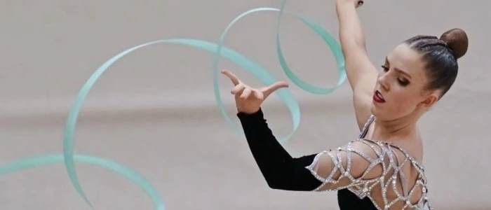Eine junge Frau trägt einen schwarz-silbernen Glitzerturnanzug und bringt führt eine Übung mit einem hellblauen Gymnastikband aus