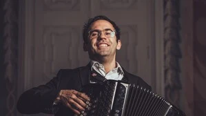 Junger Mann mit Brille hält ein Akkordeon, er lächelt, der Hintergrund ist dunkel, eine geschlossene Tür ist zu sehen