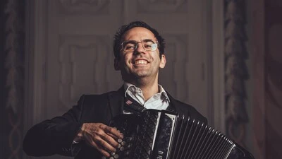 Junger Mann mit Brille hält ein Akkordeon, er lächelt, der Hintergrund ist dunkel, eine geschlossene Tür ist zu sehen