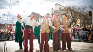 Eine Gruppe jugendlicher, verkleideter Frauen in roten Hosen und beigen Shirts geht über eine Straße, die Sonne scheint, im Hintergrund sind Absperrgitter zu sehen, zahlreiche Menschen sind dahinter zu sehen