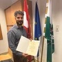 Ein junger Mannin Hemd und Lederhose steht mit einer Urkunde vor der österreichischen, steirischen und EU-Flagge