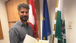 Ein junger Mannin Hemd und Lederhose steht mit einer Urkunde vor der österreichischen, steirischen und EU-Flagge