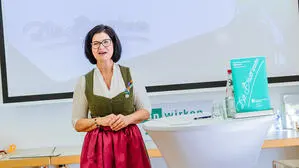 Frau mit kurzem, schwarzem Haar steht in einem grünen Dirndl mit roter Schürze vor einem digitalen Whiteboard an einem weißen Stehtisch