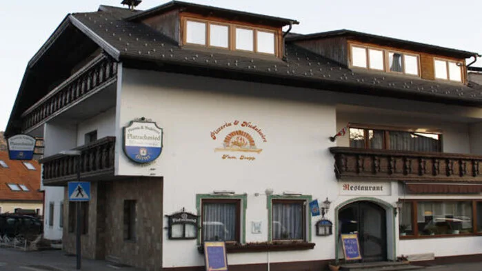Das Foto zeigt das Gasthaus in Guttaring in einer Außenansicht. Es hat dunkle Fenster und einen dunklen Holzbalkon, das Haus selbst ist weiß verputzt und hat drei Geschosse.