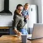 Busy mom works at home with an active toddler on her arms. Mother in glasses is watching on laptop and writing notes in the kitchen while holding kid