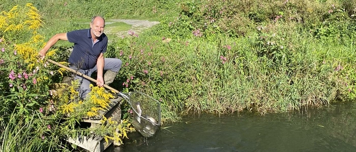 Ein Fischzüchter steht mit einem Kescher an einem seiner Teiche und lächelt in die Kamera.
