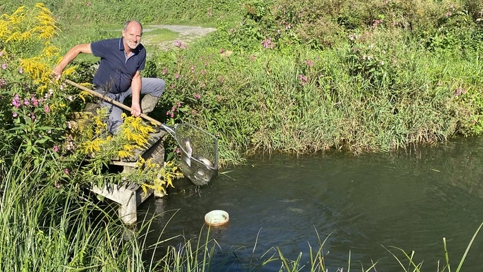 Ein Fischzüchter steht mit einem Kescher an einem seiner Teiche und lächelt in die Kamera.
