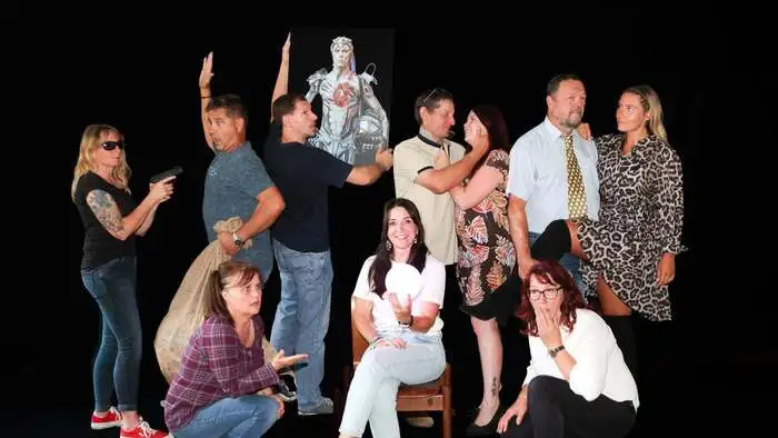 Das Bild zeigt mehrere Darsteller der Theatergruppe St. Ulrich, die alle in verschiedenen Posen stehen oder hocken. In der Mitte sitzt auf einem Stuhl Obfrau Petra Hatzel.