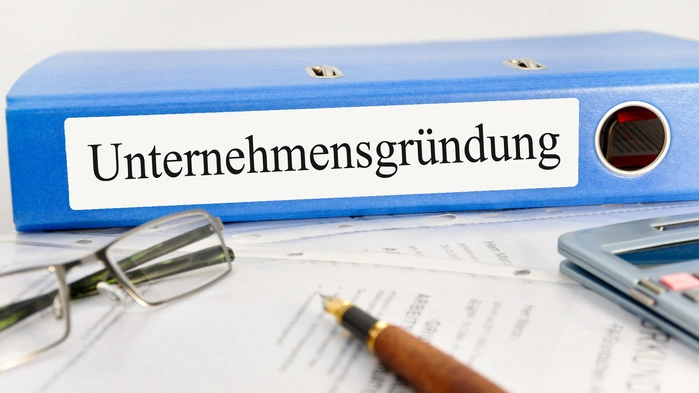 Ein Ordner mit der Aufschrift „Unternehmensgründung“ liegt auf einem Tisch neben einem Taschenrechner und ein Papier mit Stift 