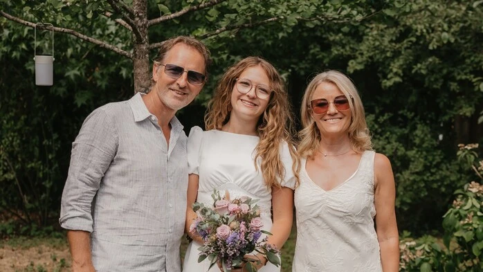 Andy und Kelly Kainz mit ihrer Tochter Grace Kelly. Sie stehen in einer Reihe. Andy trägt ein helles legeres Hemd und eine passende Hose, Kelly ein weißes, ärmelloses bodenlanges Kleid mit V-Ausschnitt. Grace trägt ein kurzes weißes Etuikleid und hält einen Blumenstrauß in der Hand.