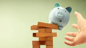 Piggy bank suddenly falls off unstable Jenga block, hands protecting it, symbolizing fragile financial stability and economic uncertainty, need for caution