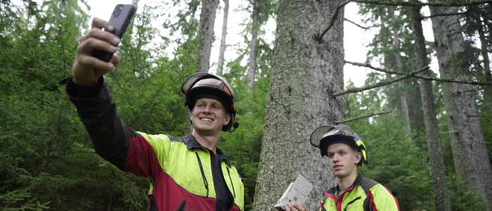 Zwei Männer stehen in Forstmontur in einem Wald vor einem Baum und machen ein Selfie